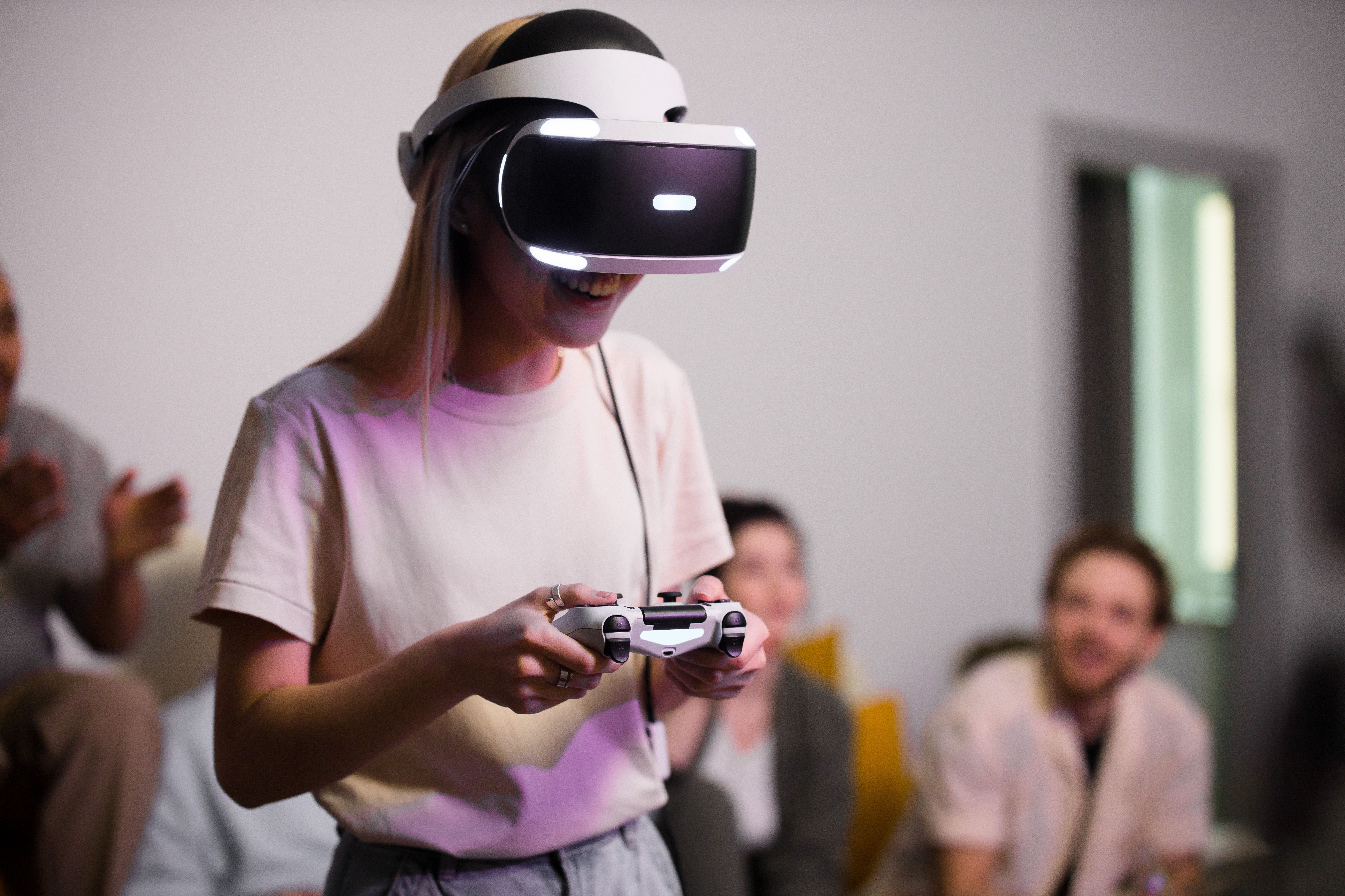 A person wearing a virtual reality headset smiles while holding a game controller, surrounded by friends who are engaged and watching her play.