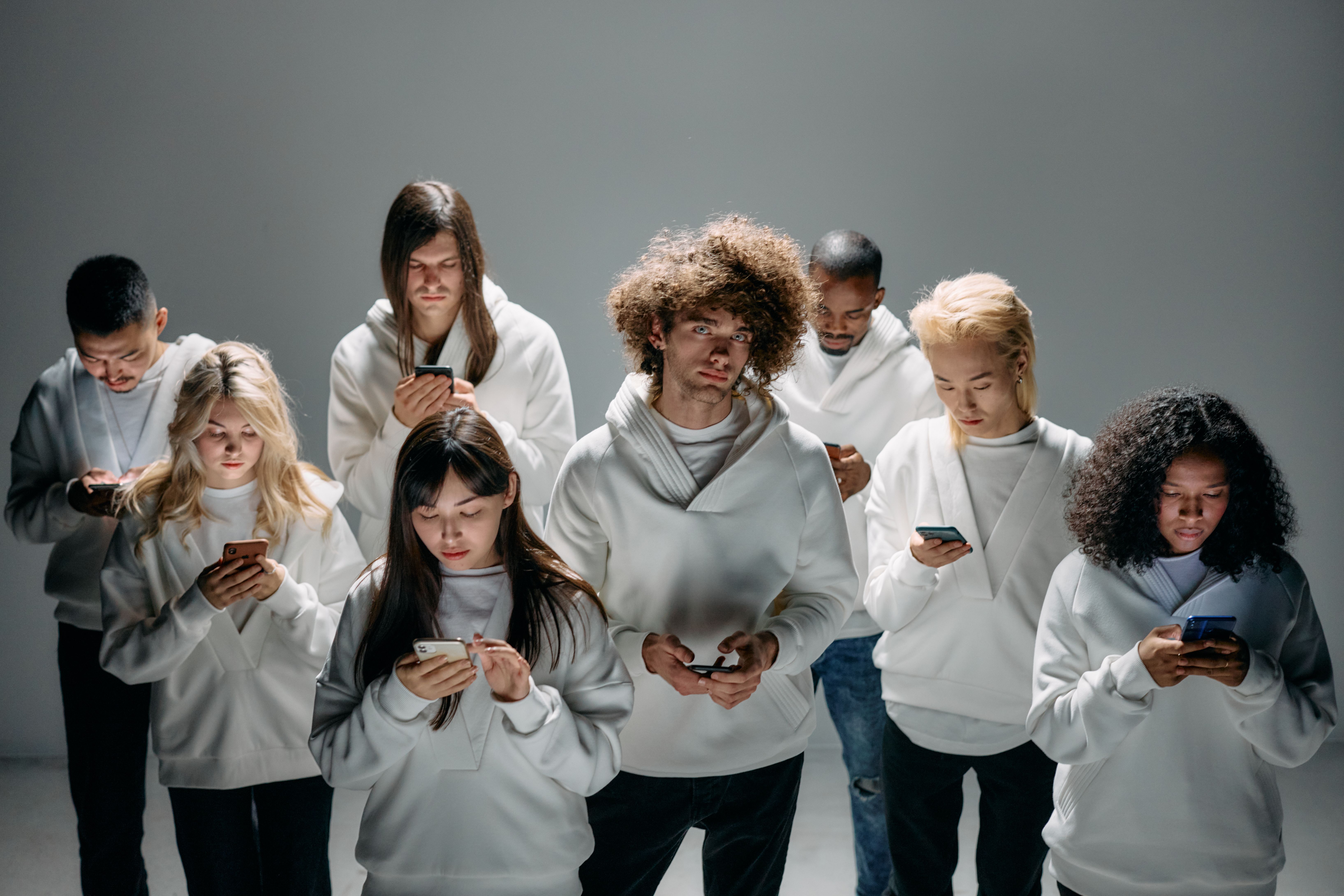 A diverse group of eight individuals wearing white hoodies, focused on their smartphones in a dimly lit studio setting.